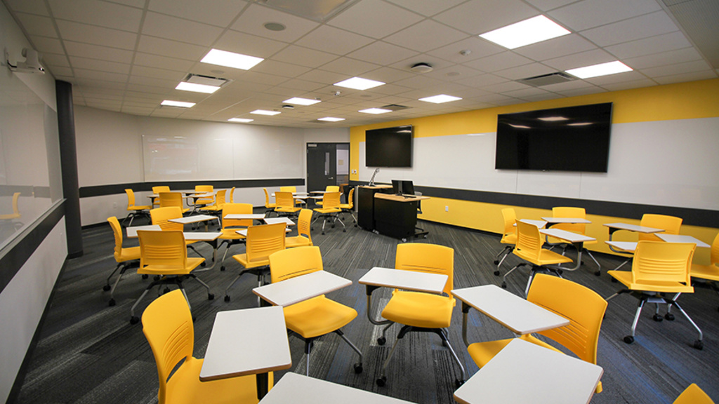 Desks are arranged in circles in Lindquist Center room S112, with large monitors on one wall and another wall covered with a large whiteboard.